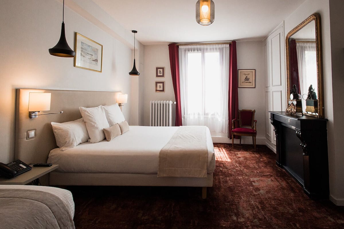 A hotel room with a bed, bedside lamps, a desk with a phone, a chair, a dresser, and a large mirror.