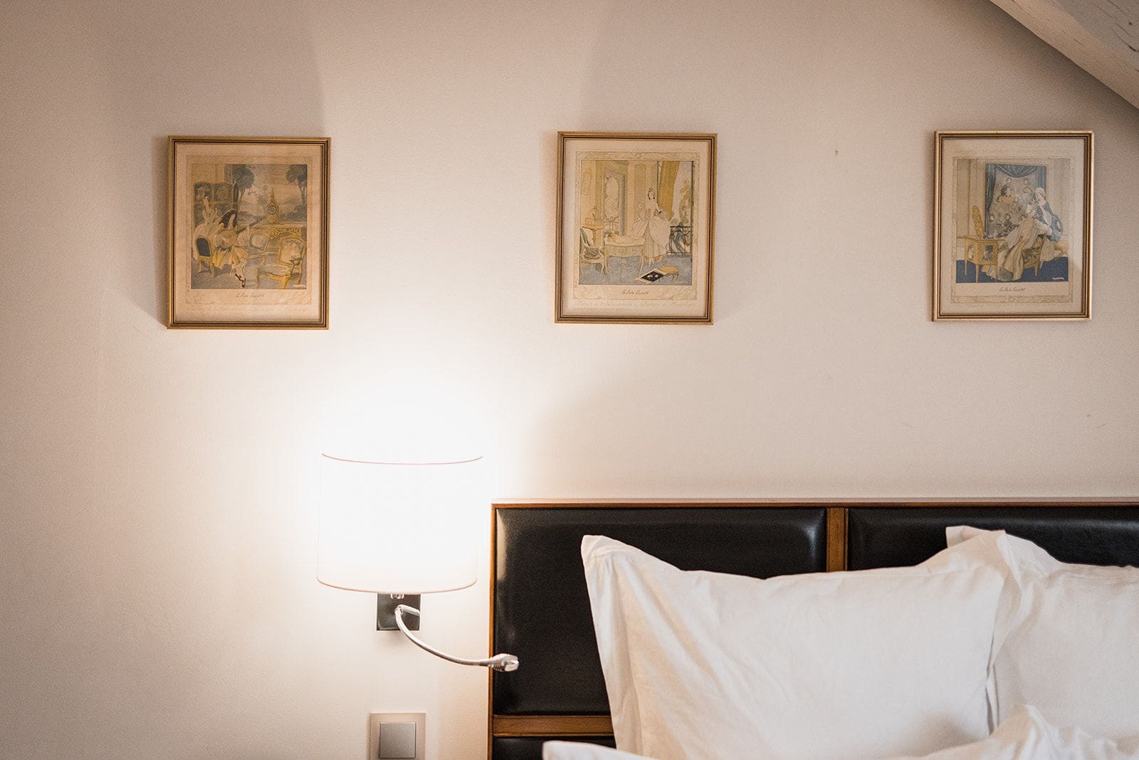 A hotel room with a bed, a nightstand, and a wall-mounted lamp. There are three framed pictures on the wall.