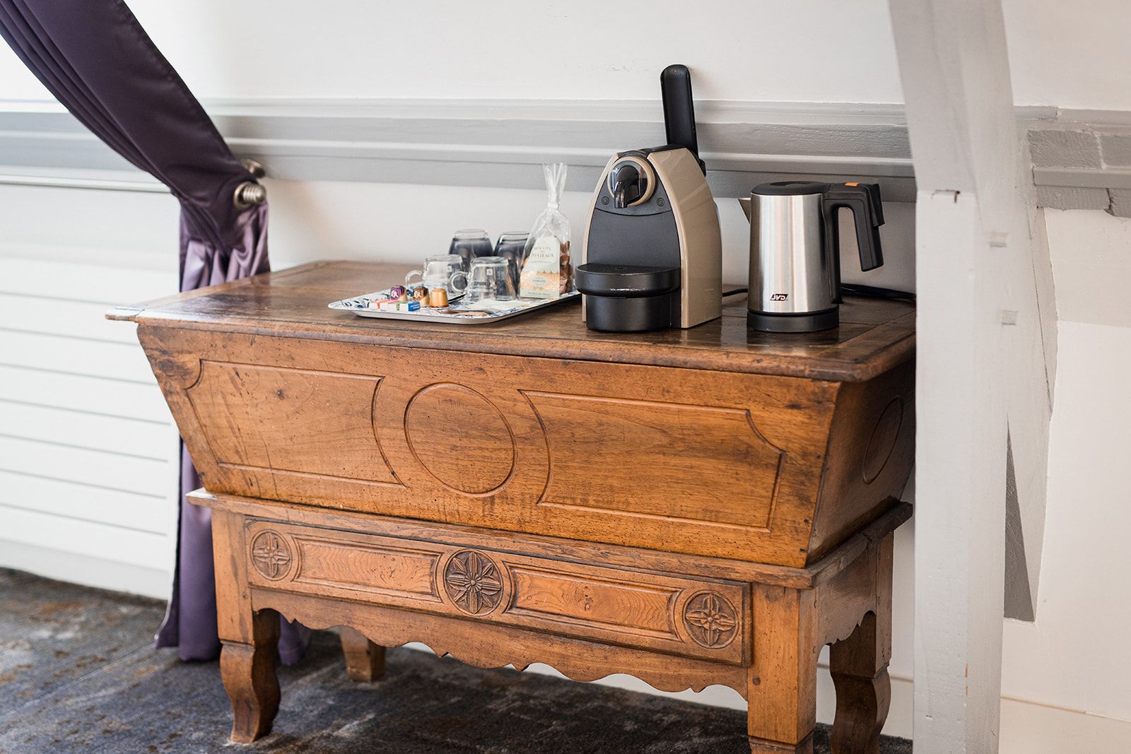 A wooden chest of drawers with a coffee maker, a kettle, and a tray with mugs and a bottle on top.