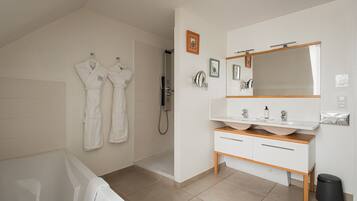 Une salle de bain moderne avec une baignoire, une douche, un meuble-lavabo avec un miroir et deux peignoirs blancs accrochés au mur.
