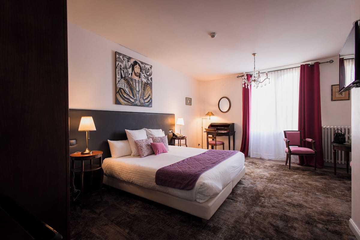 A hotel room with a bed, a desk, a chair, a lamp, a painting, and a television.