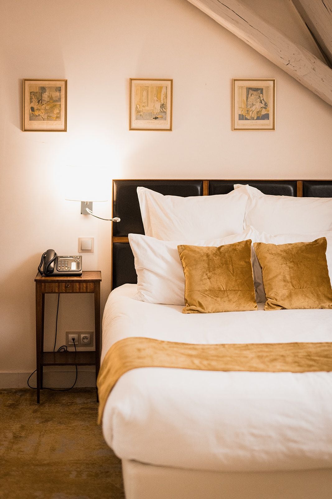 A hotel room with a bed, a bedside table with a telephone, and three framed pictures on the wall.