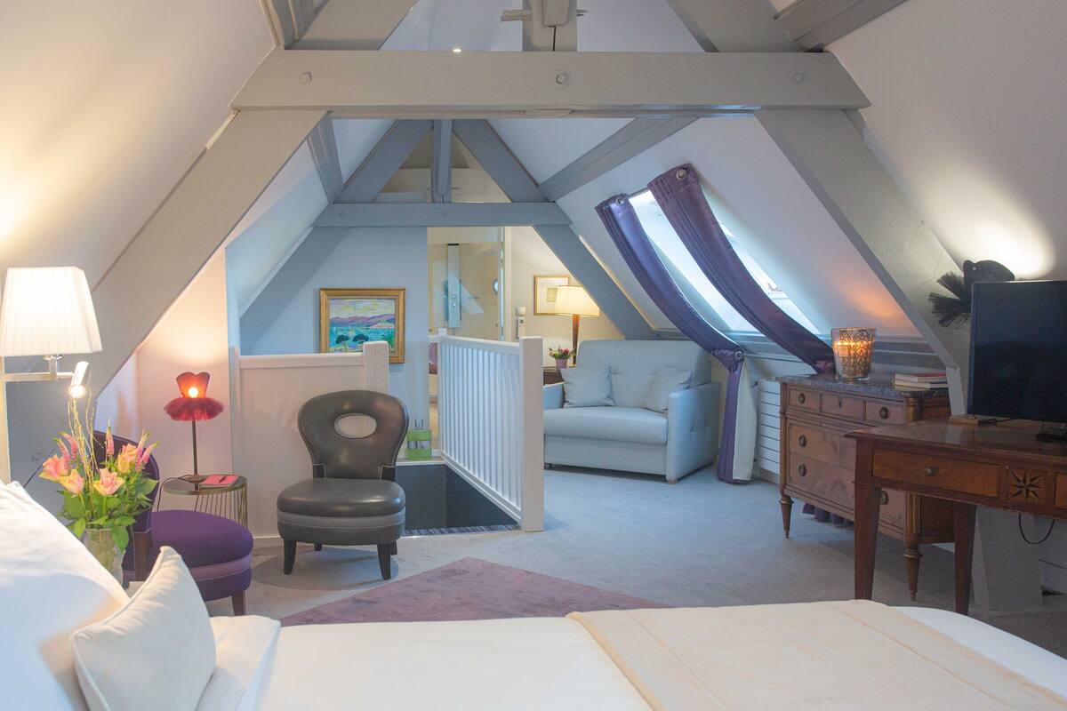 A bedroom with a sloped ceiling, a bed, a chair, a dresser, and a television.