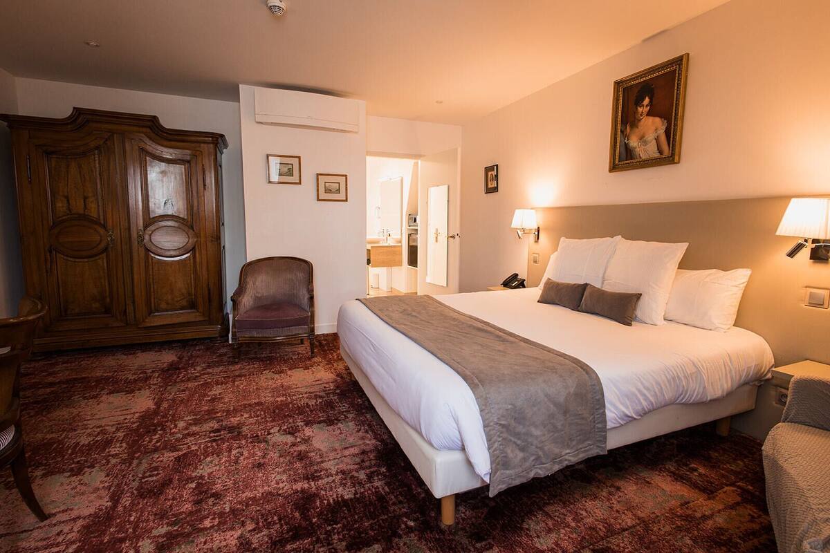 A hotel room with a large bed, a wooden wardrobe, a chair, and a red carpet.