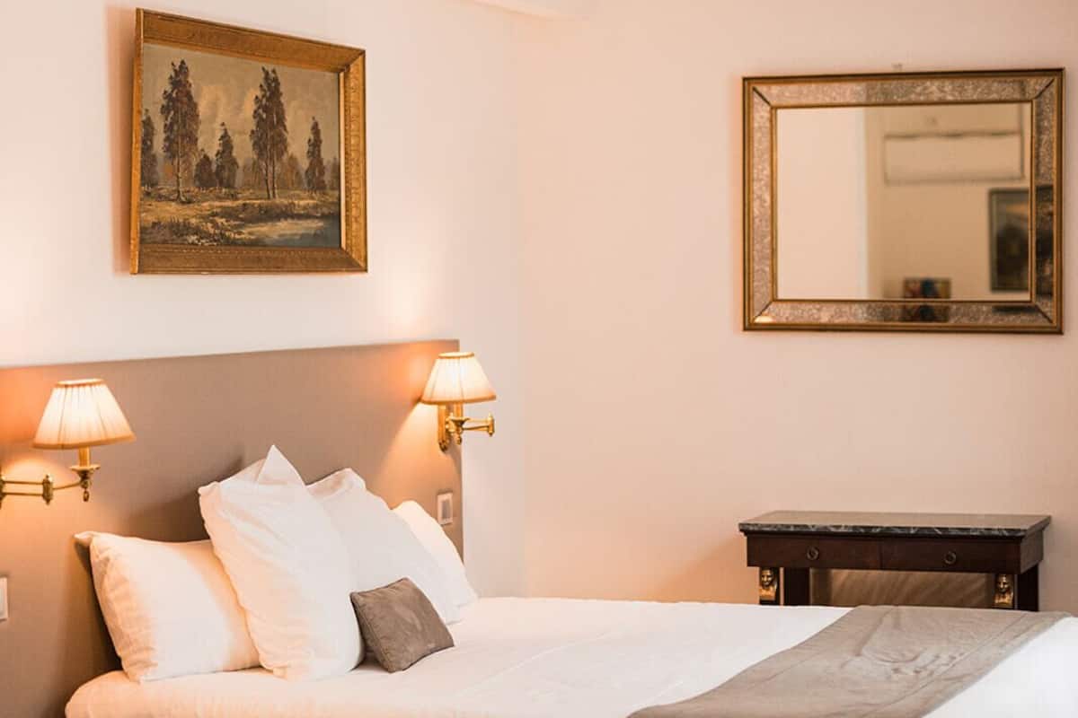 A hotel room with a bed, two wall-mounted lamps, a painting, a mirror, and a small table.