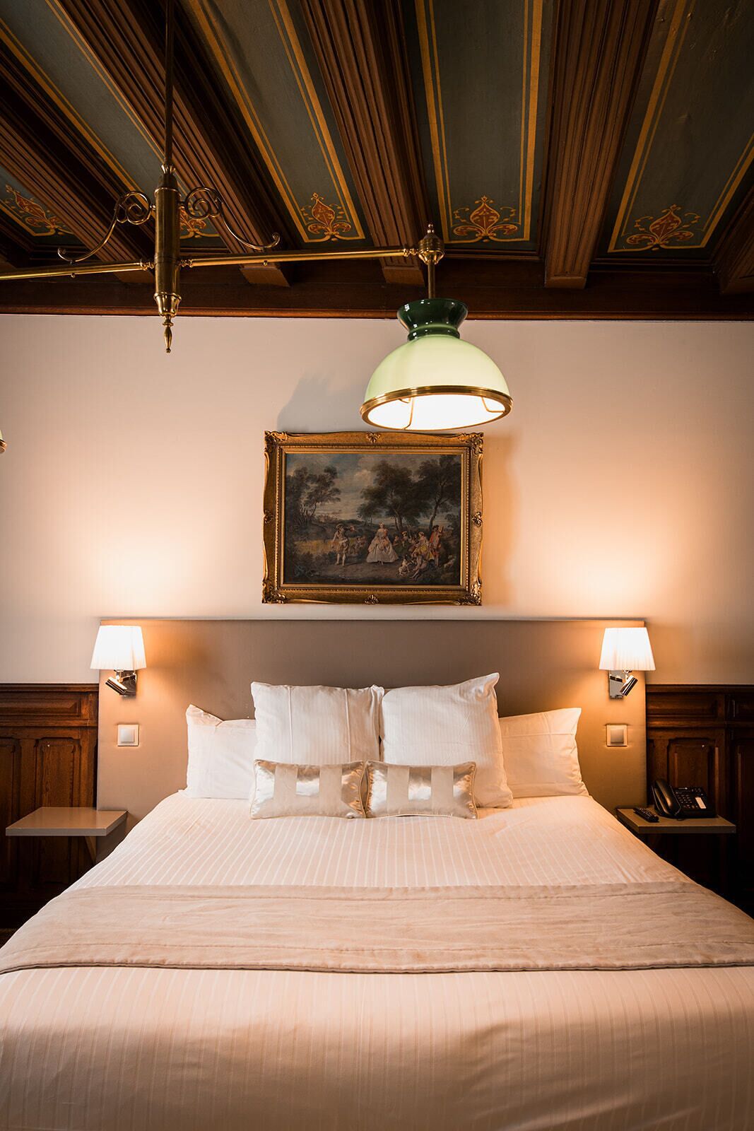 A hotel room with a large bed, two bedside lamps, a painting above the bed, and a chandelier.
