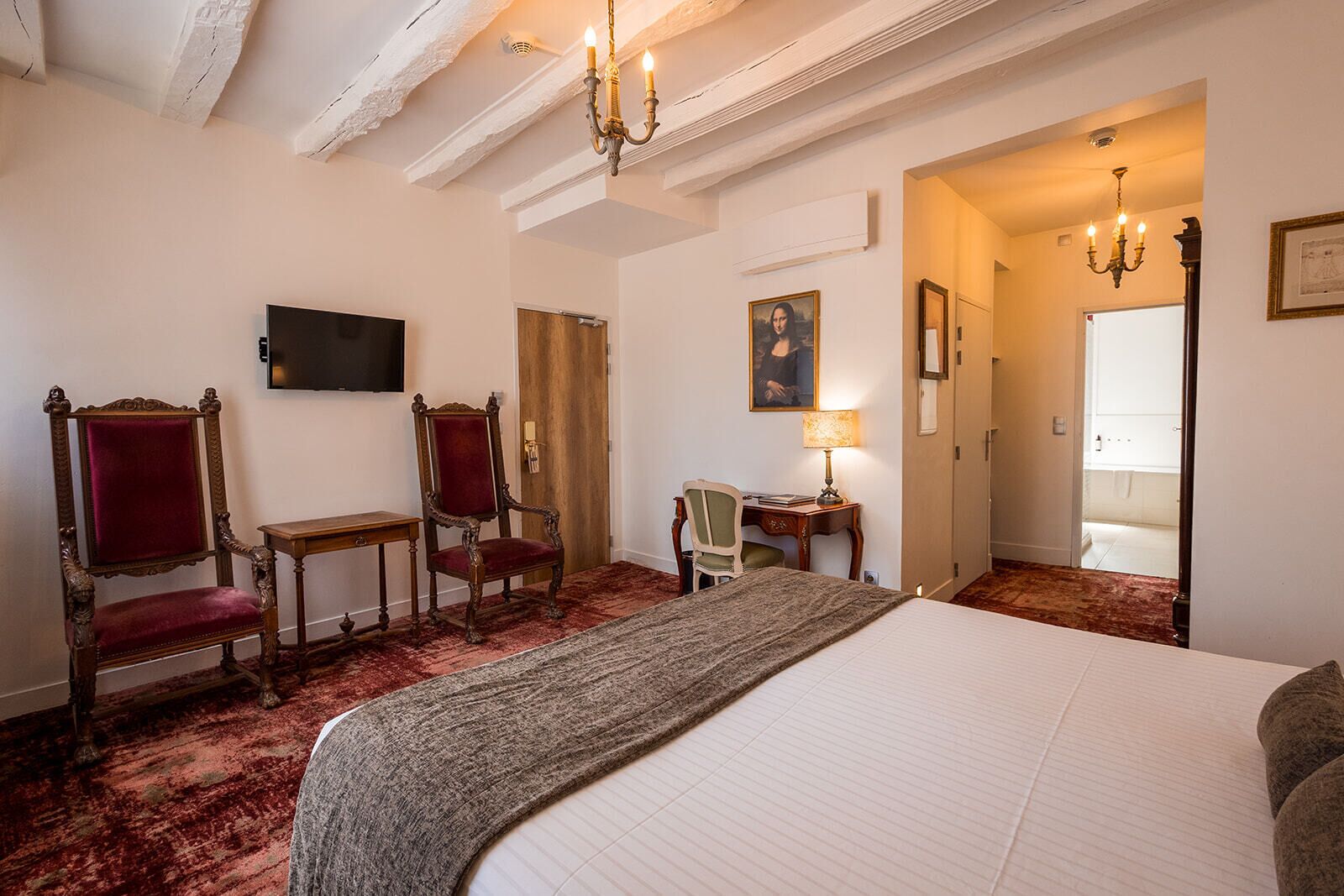 A hotel room with a large bed, two ornate chairs, a desk with a lamp, a television, and a painting on the wall.