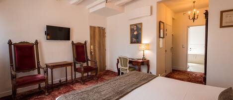 A hotel room with a large bed, two ornate chairs, a desk with a lamp, a television, and a painting on the wall.