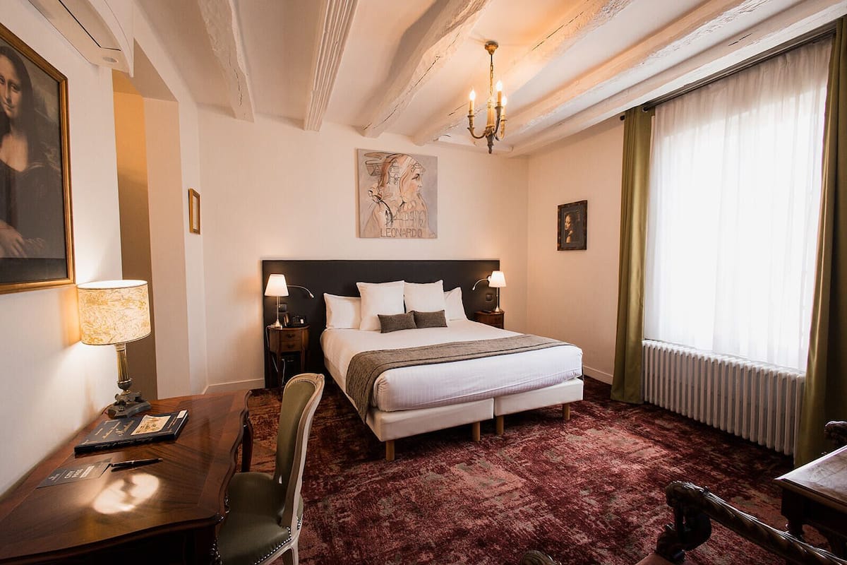 A hotel room with a large bed, a desk with a lamp, a chair, a painting on the wall, and a chandelier.