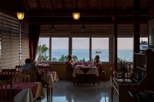 A restaurant with tables and chairs, large windows with a view of the sea and a ship, and hanging light fixtures.