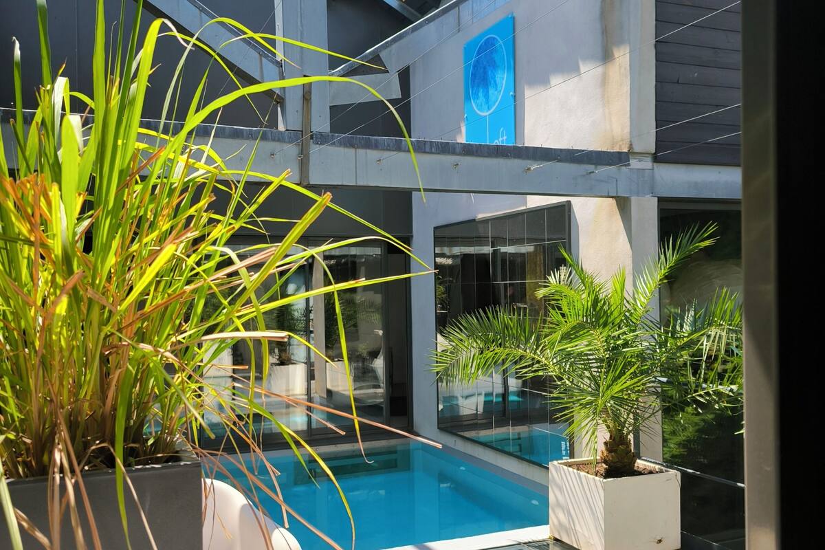 A modern outdoor pool area with glass walls, potted plants, and a clear blue sky.