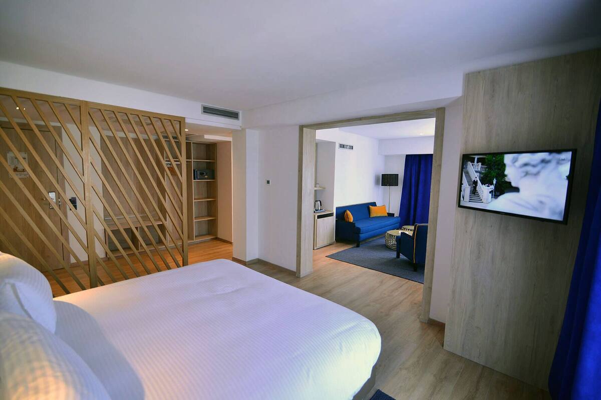 Habitación de hotel con cama, escritorio, silla, televisor y vista al pasillo y otra habitación.