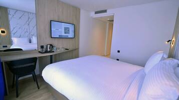 A hotel room with a bed, a desk, a chair, a TV mounted on the wall, and a QR code on the screen.