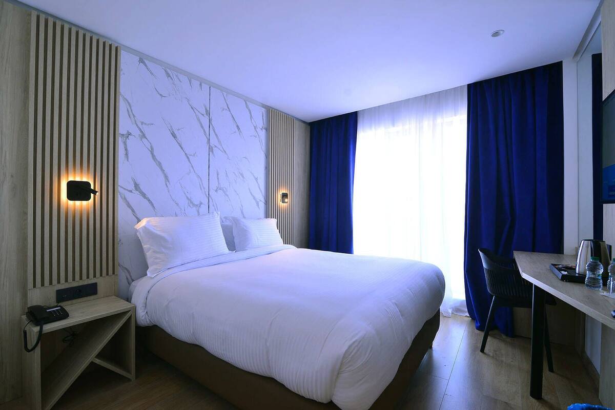 Una habitación de hotel moderna con una cama grande, un escritorio y una silla. Hay dos luces fijadas a la pared y una pared con diseño de mármol.