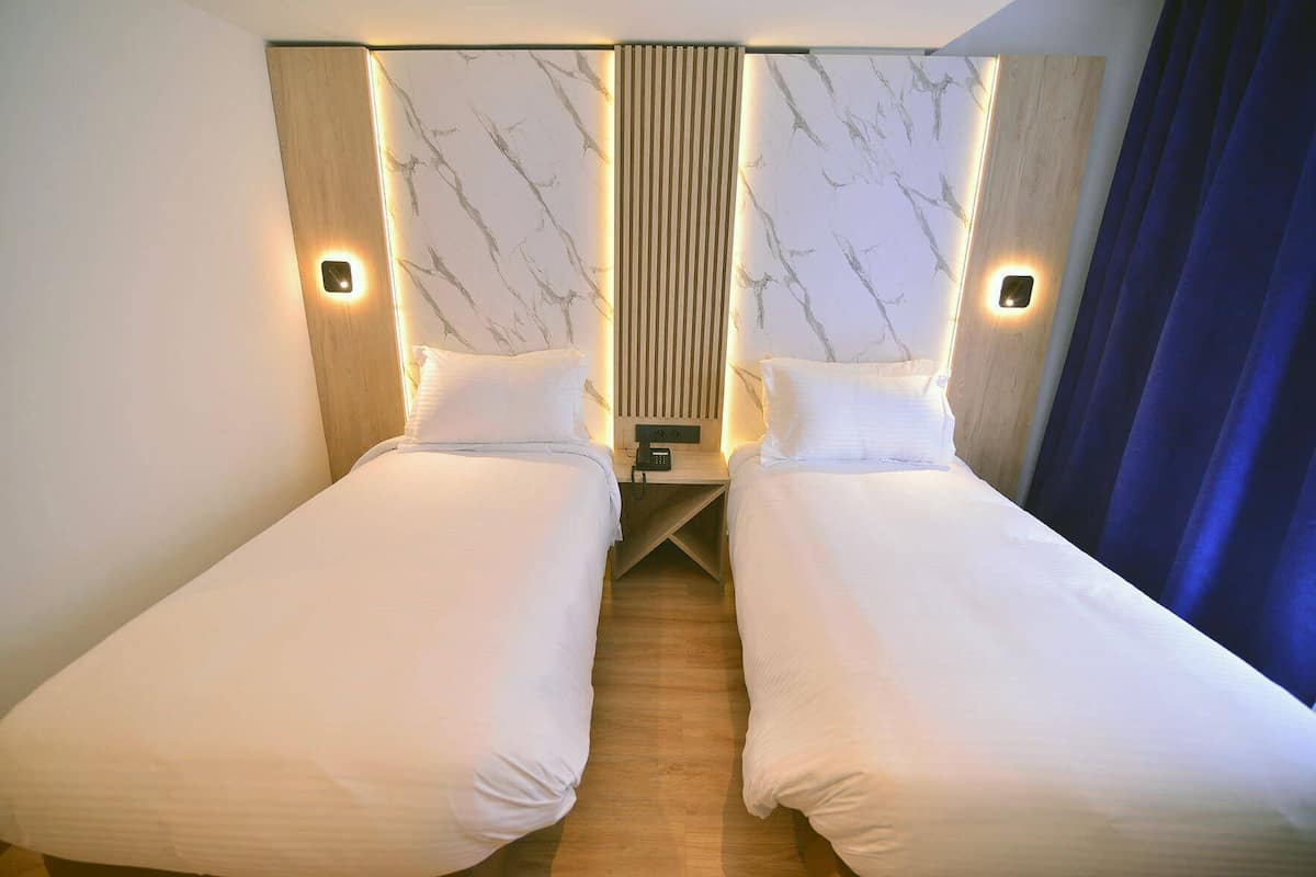 Habitación de hotel con dos camas individuales, cabecera de madera, paneles de pared con diseño de mármol, mesitas de noche con lámparas y cortina azul.