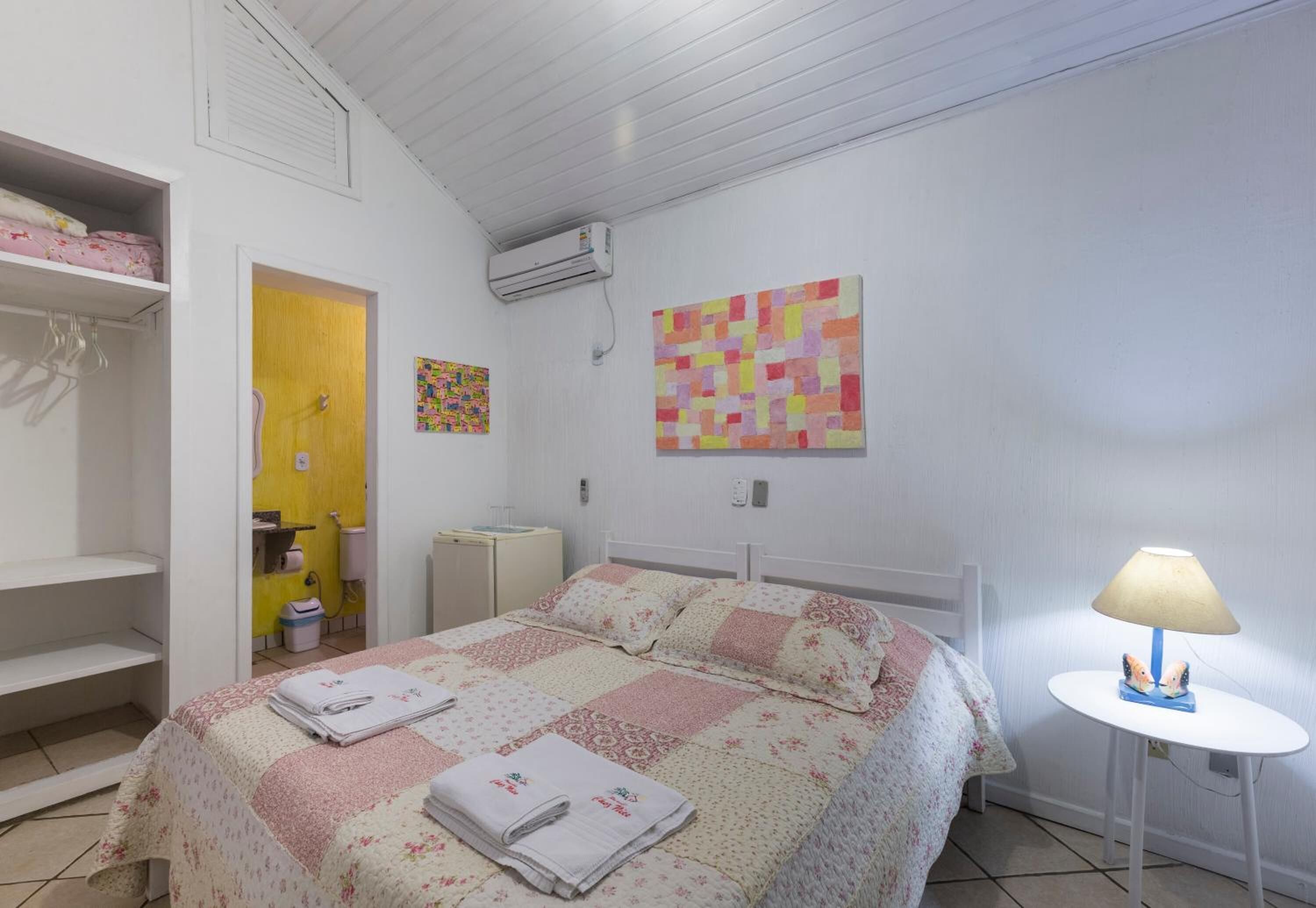 A bedroom with a bed, a bedside table with a lamp, a wardrobe, a small table with a lamp, and a wall with a colorful artwork.
