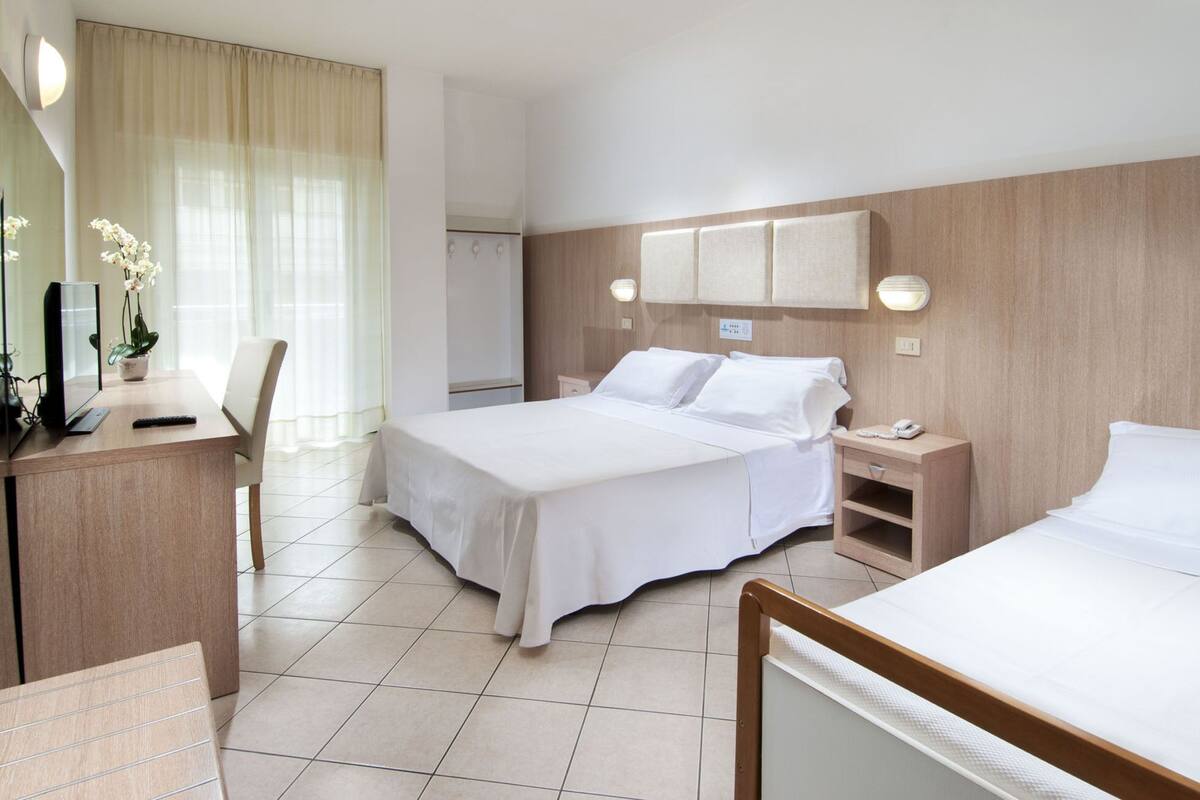 A hotel room with a bed, bedside tables, a desk with a chair, a mirror, and a potted plant.