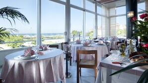 A restaurant with tables set for dining, large windows offering a view of the sea, and a potted plant on the left. - Hotel Cormoran (Cattolica)