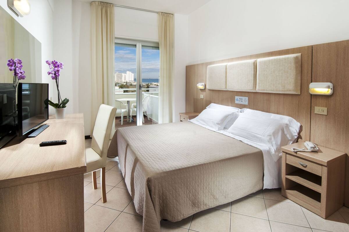 A hotel room with a bed, a desk with a TV, a chair, a nightstand, and a view of the city.