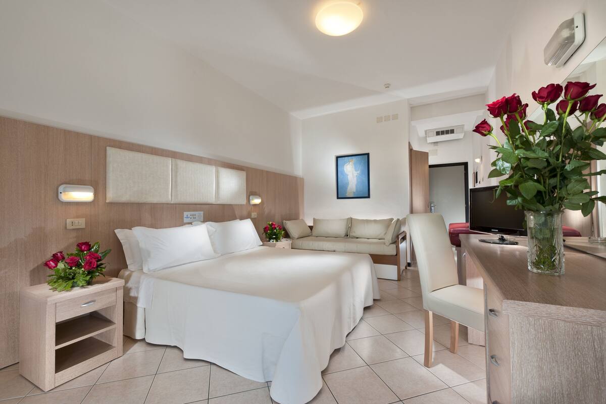 A modern hotel room with a large bed, a sofa, a TV, and a dining area.
