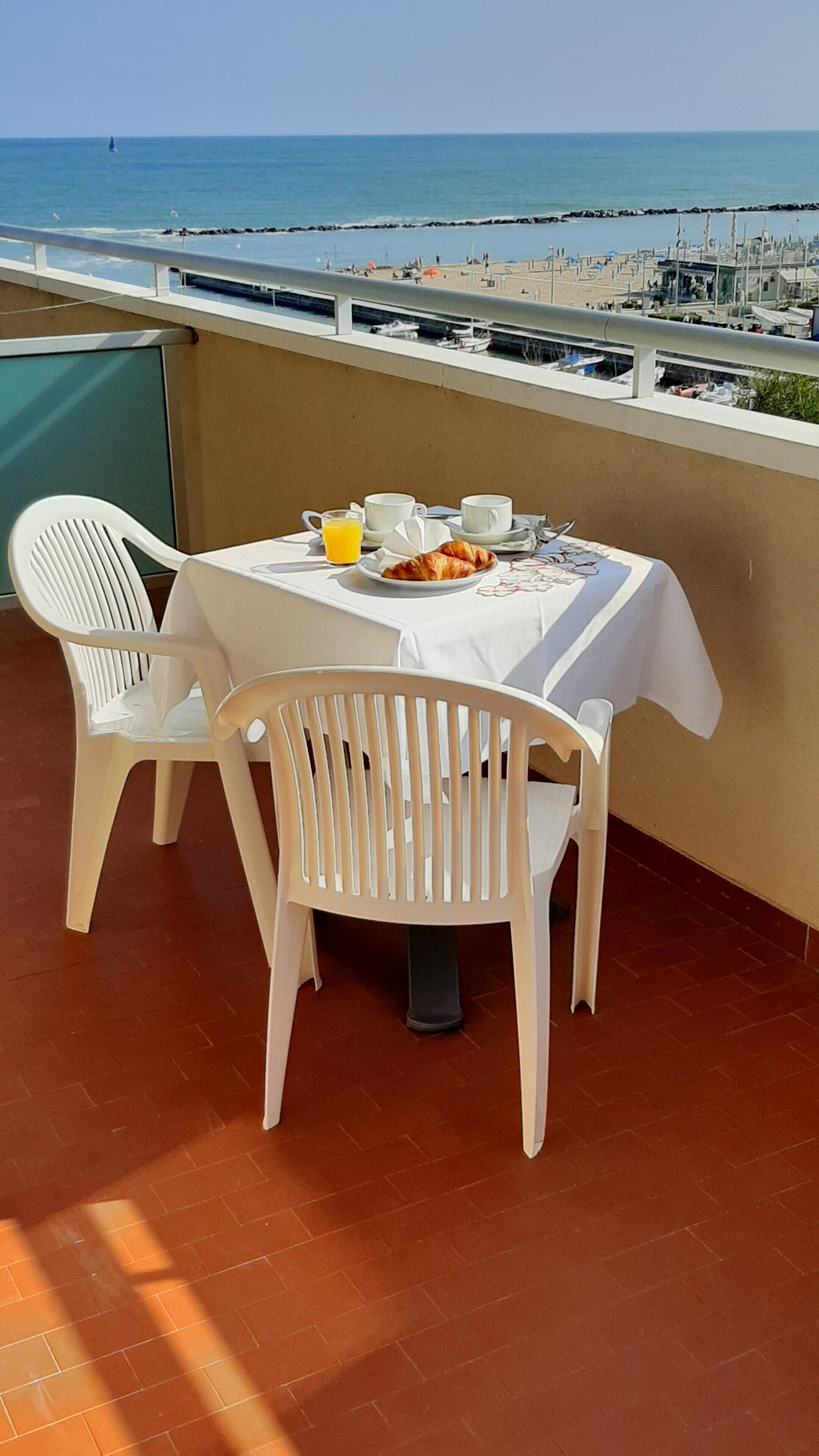 Mesa dispuesta para el desayuno, dos sillas blancas y vista al mar y barcos.