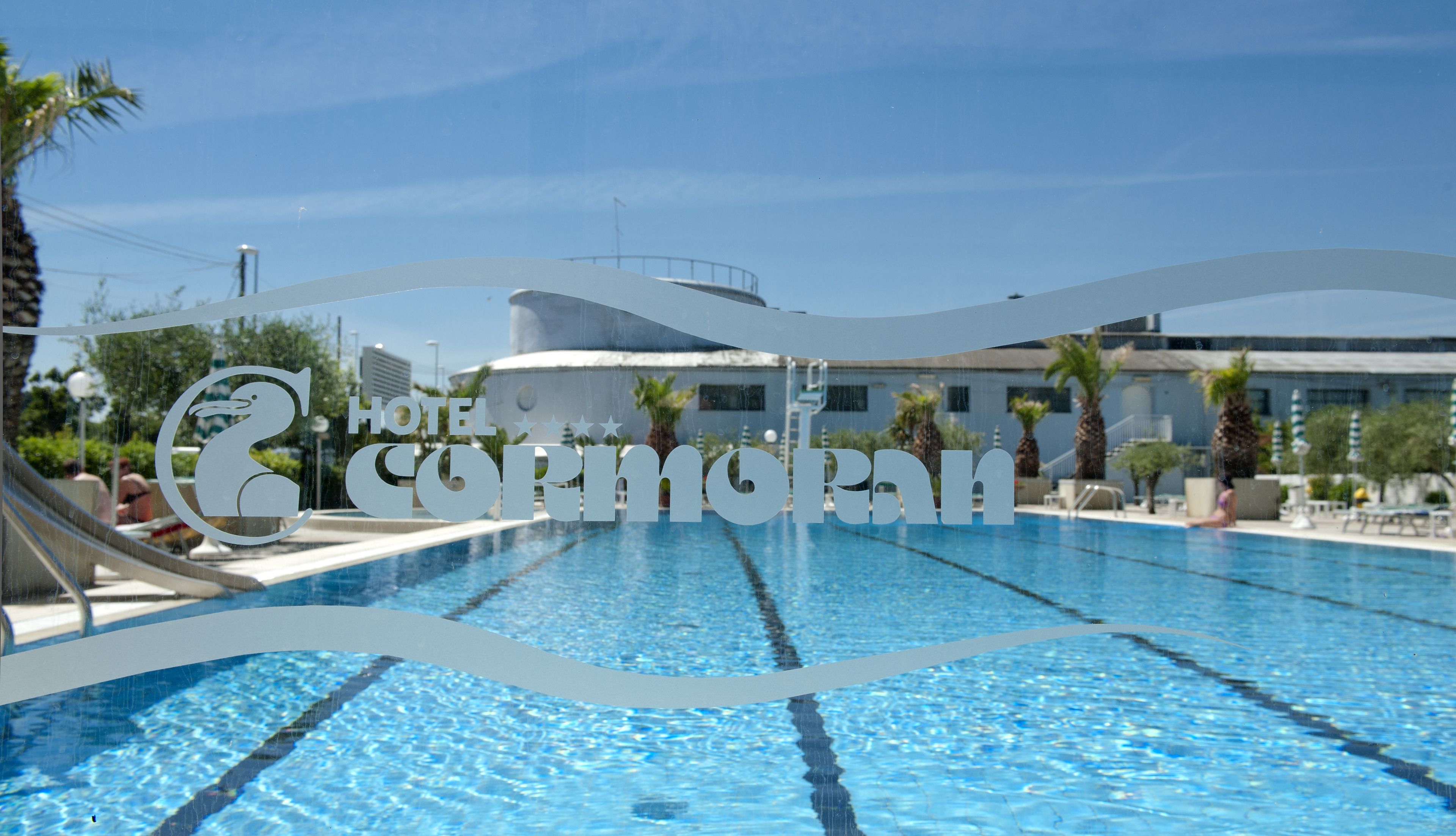 A swimming pool with clear blue water, surrounded by palm trees and a hotel building in the background.