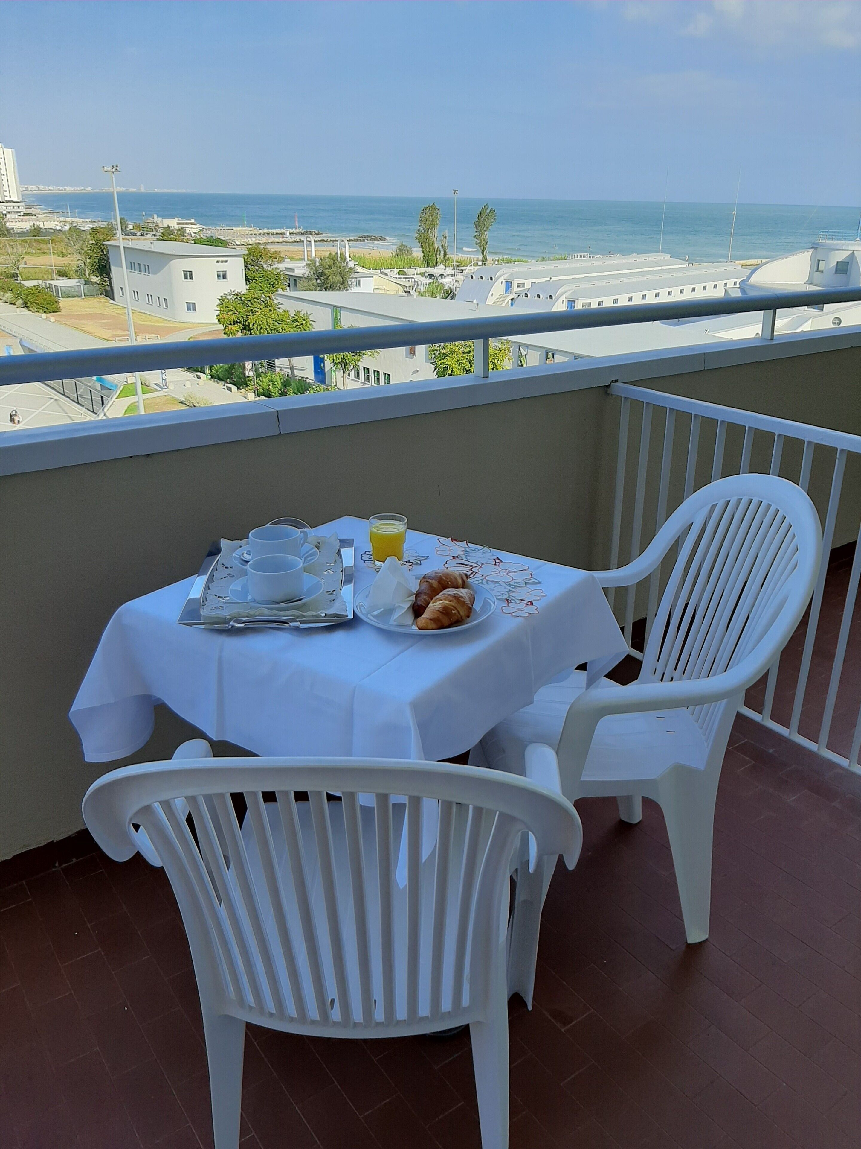 Un balcón con una mesa lista para dos personas, sillas de plástico blanco y vista al mar y edificios al fondo.