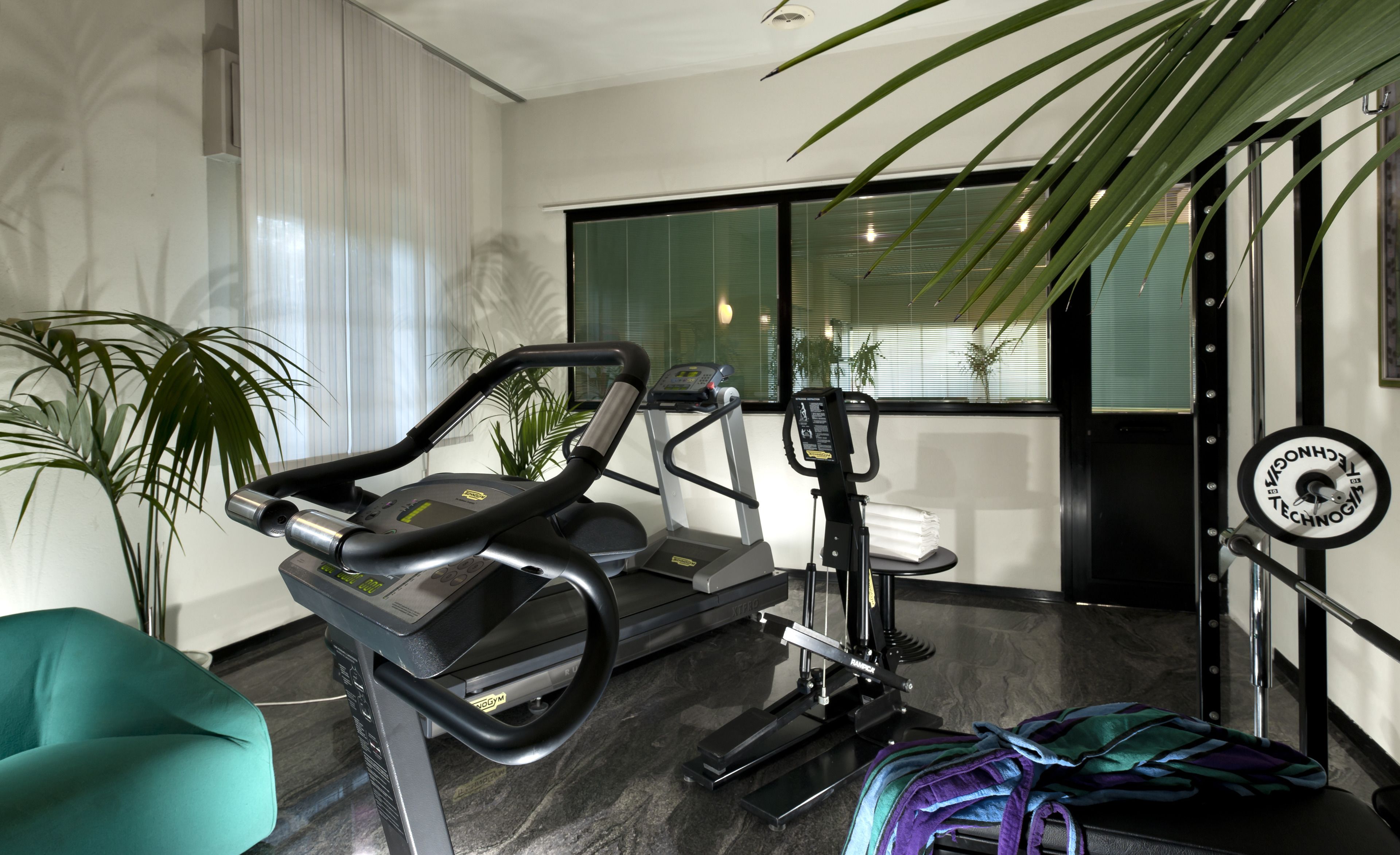 Une salle de sport moderne équipée d’appareils de cardio, d’un fauteuil de couleur turquoise et d’une grande fenêtre avec des stores.