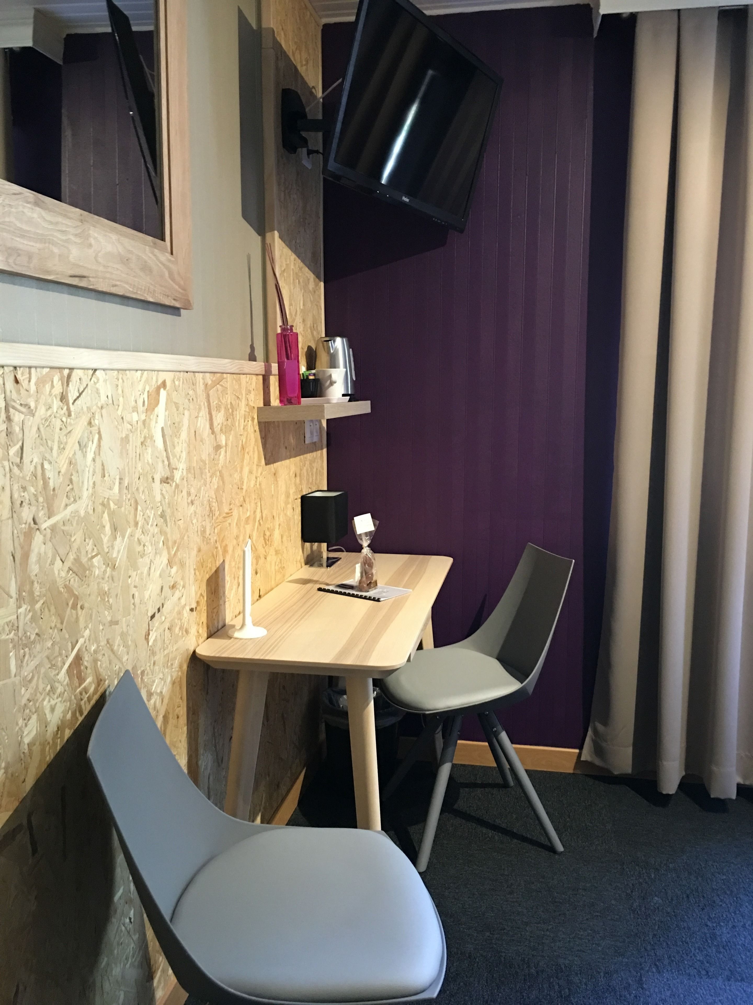 Hotellihuone, jossa on puinen seinä, puisen pöydän yläpuolelle asennettu litteänäyttöinen televisio, kaksi tuolia, pieni hylly, jossa on vaaleanpunainen maljakko ja vedenkeitin, sekä violetti verho.