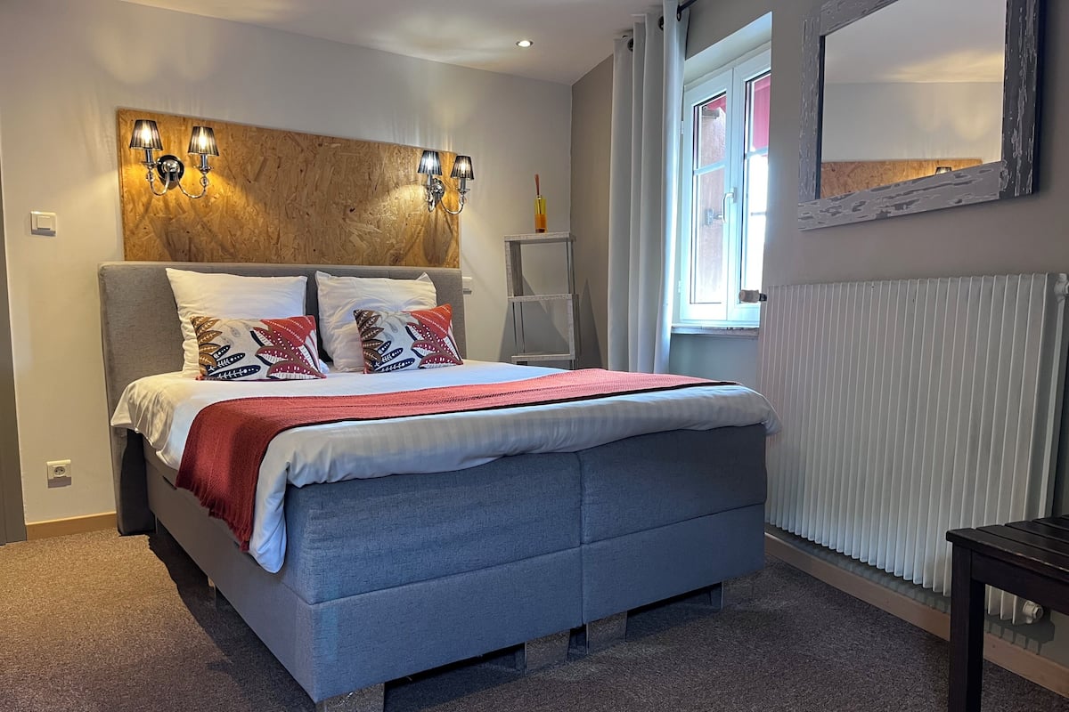 A bedroom with a bed, a headboard with wall-mounted lights, a window with curtains, and a radiator.