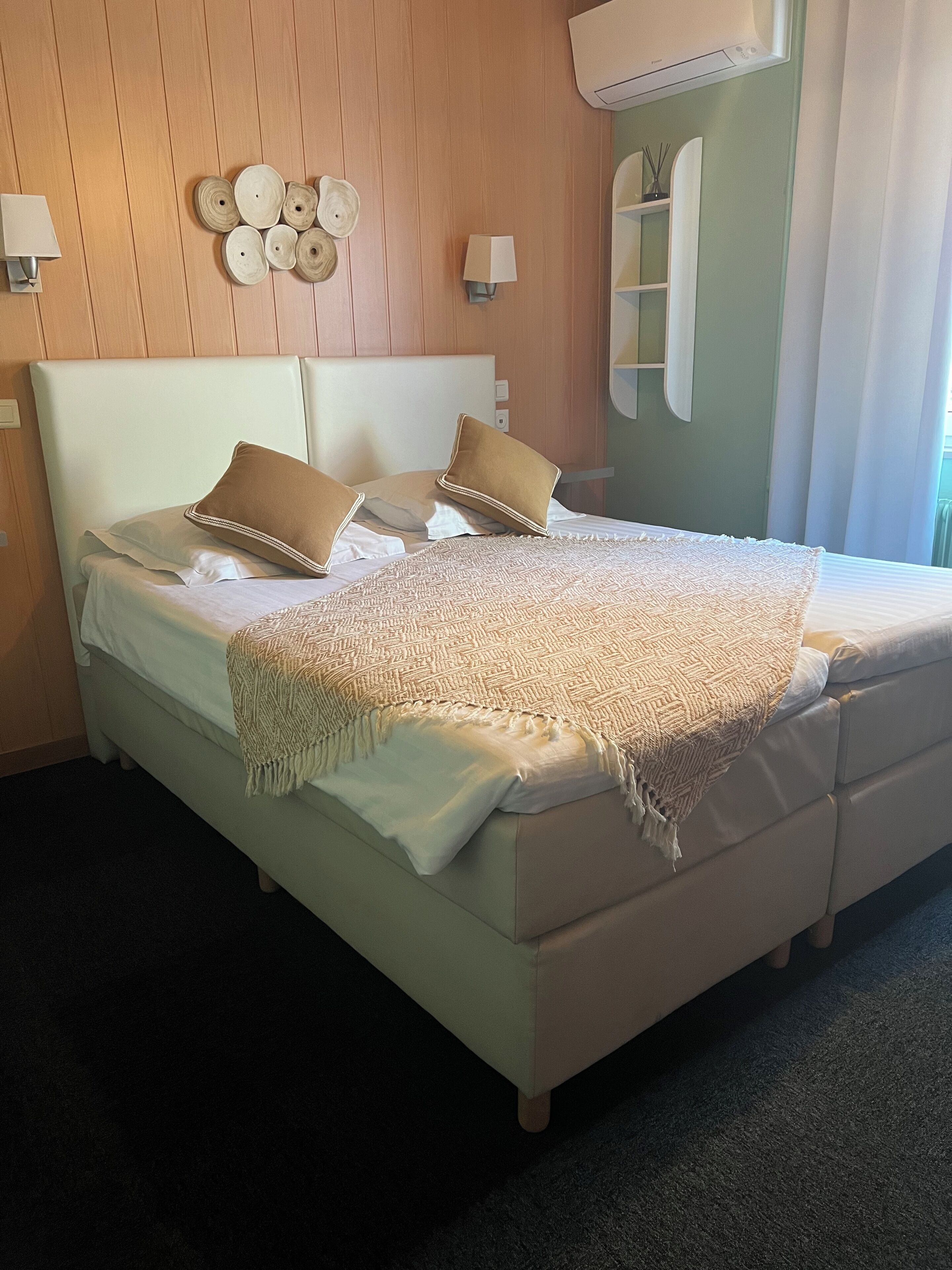 A neatly made bed with a patterned blanket and two pillows, a wall-mounted light fixture, and a mirror with a shelf above it.