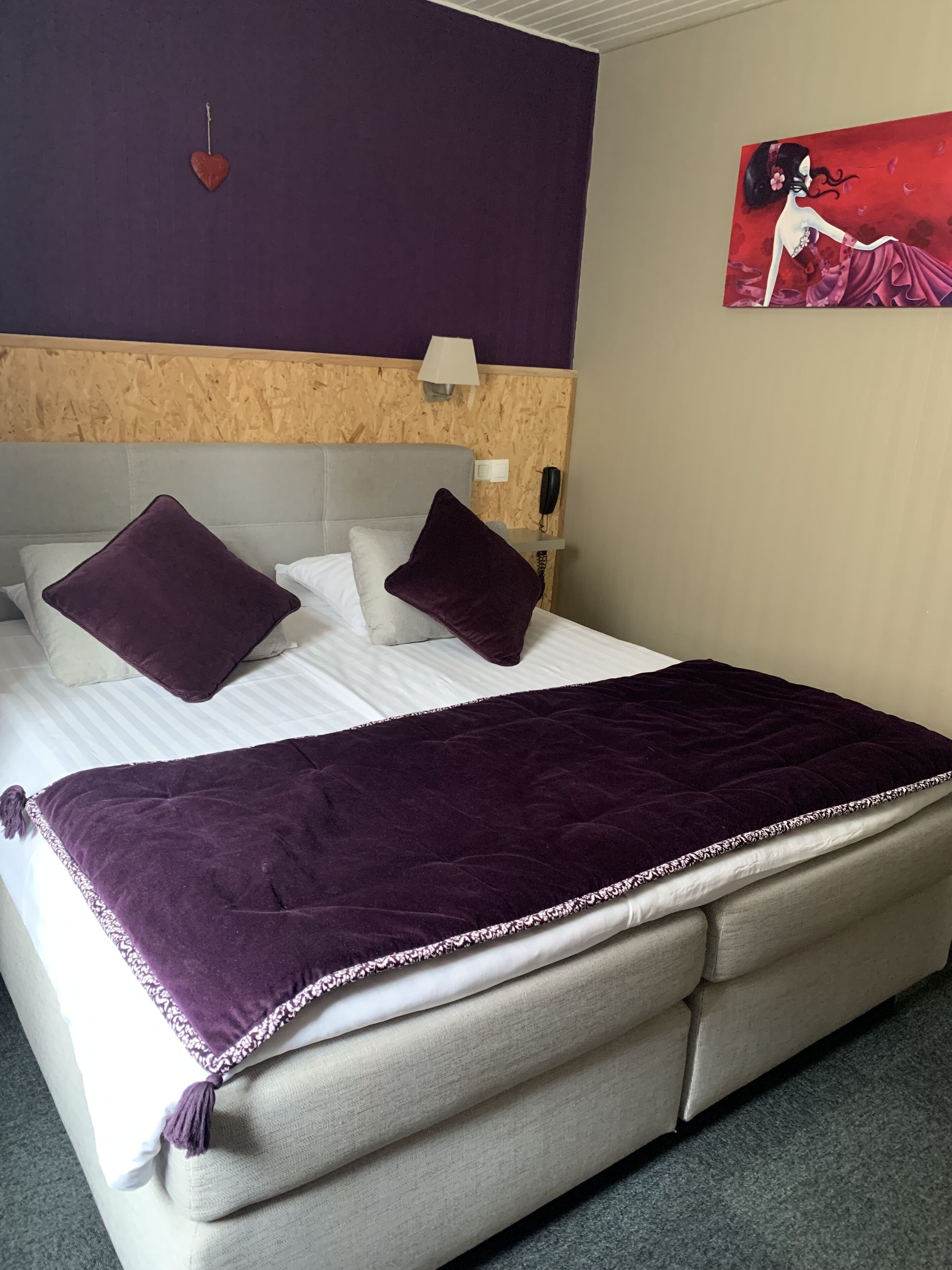A bed with a headboard, two pillows, a blanket, a wall-mounted phone, and a painting.