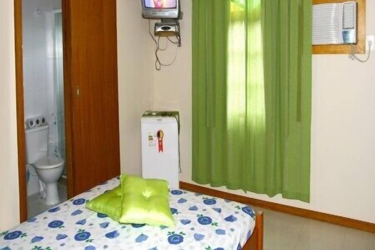 Quarto pequeno com cama, televisão fixada na parede, frigorífico e janela com cortinas verdes.