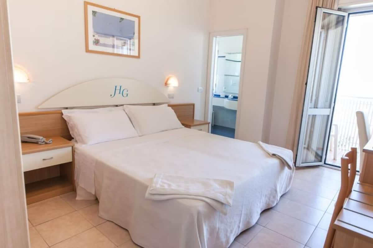 A hotel room with a bed, bedside tables, a phone, a mirror, and a view of the balcony.