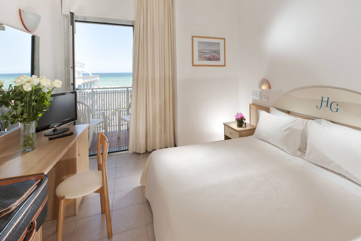 A hotel room with a bed, a desk with a TV, a chair, a nightstand with a lamp, and a view of the sea.