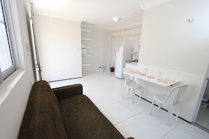 A modern living room with a brown sofa, a dining area with a white table and chairs, and a kitchen with a refrigerator and shelves. - Residencial Santa Lucia (Fortaleza)