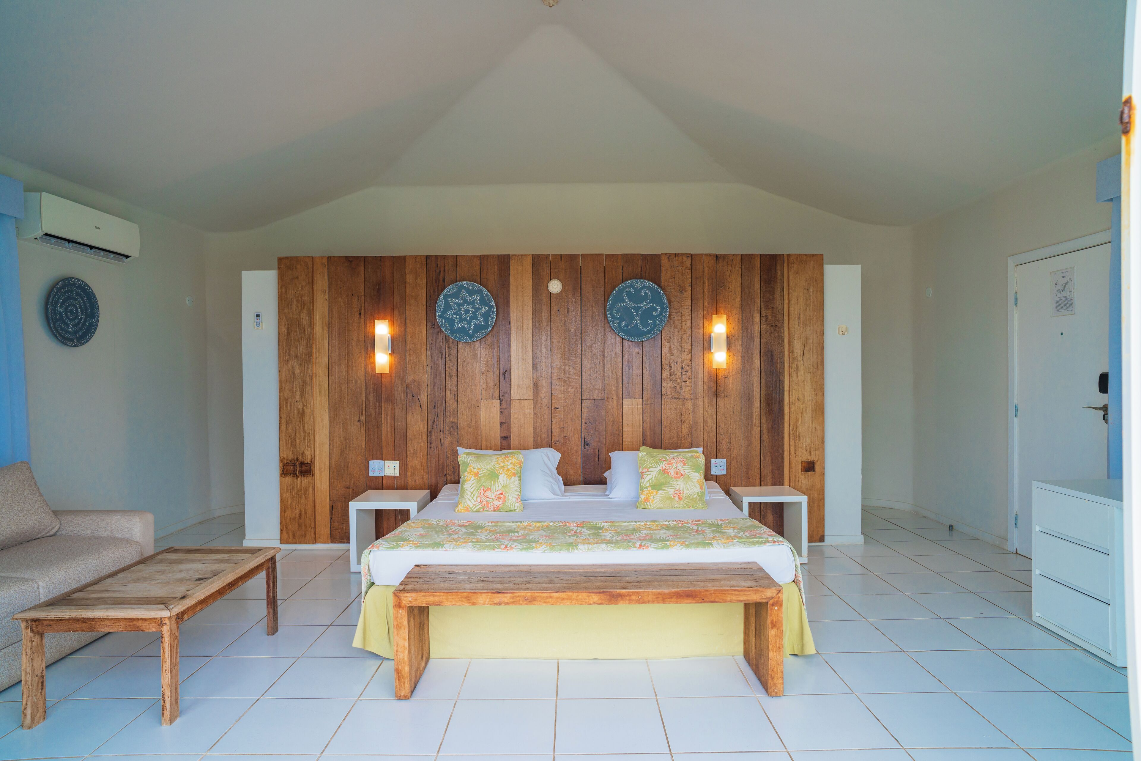 A bedroom with a wooden headboard, a bed with floral bedding, a bench, a nightstand, and a dresser.