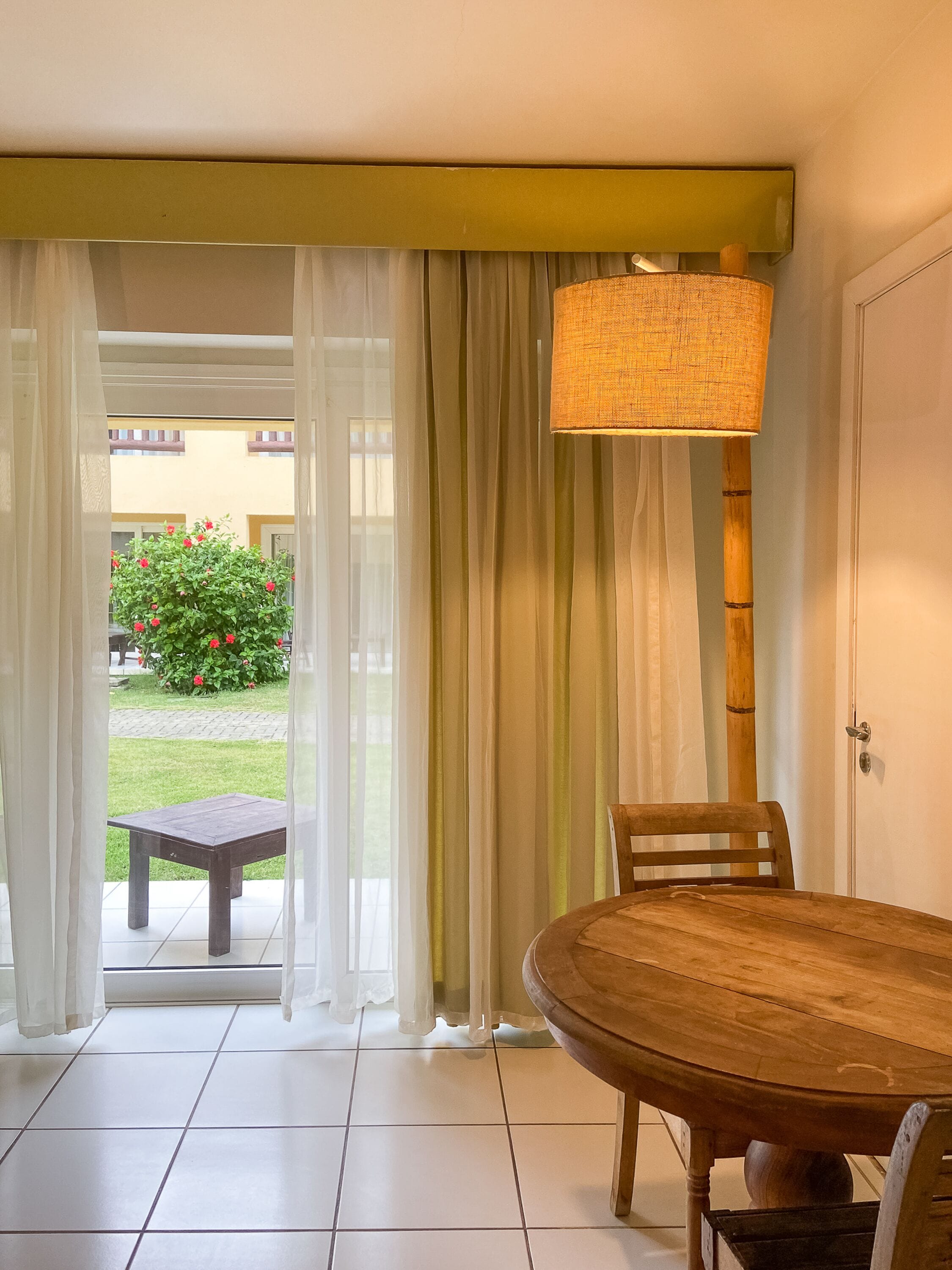 Un coin repas avec une table et des chaises en bois, une lampe suspendue et une vue sur un jardin à travers des rideaux légers.