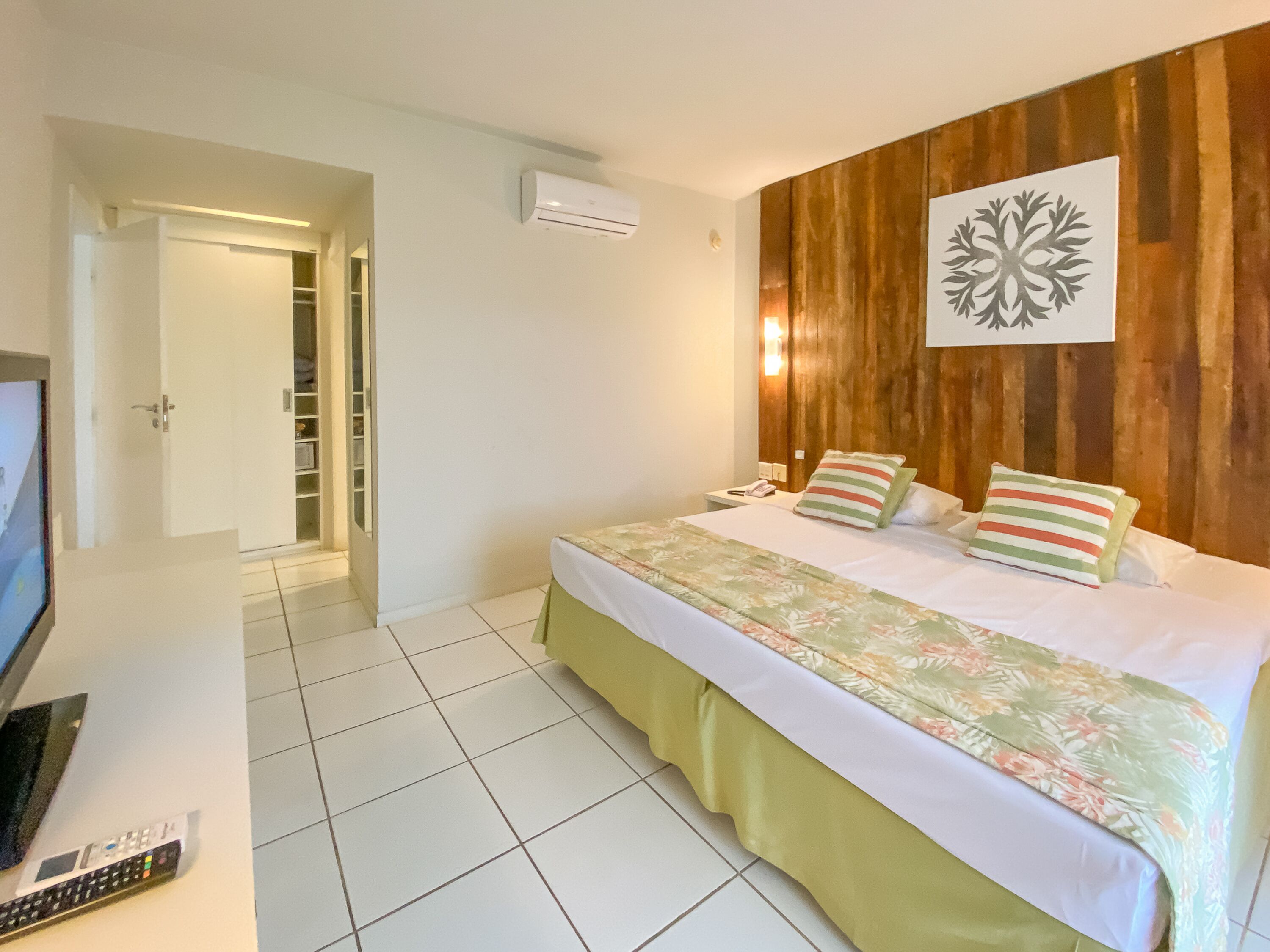Quarto de hotel com cama, televisão, controle remoto e parede de madeira com um detalhe decorativo.