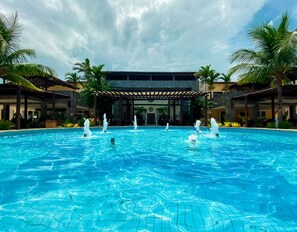 A swimming pool with multiple water jets, surrounded by palm trees and a covered area with a stone facade. - Búzios Beach Resort by WAM Experience (Búzios)