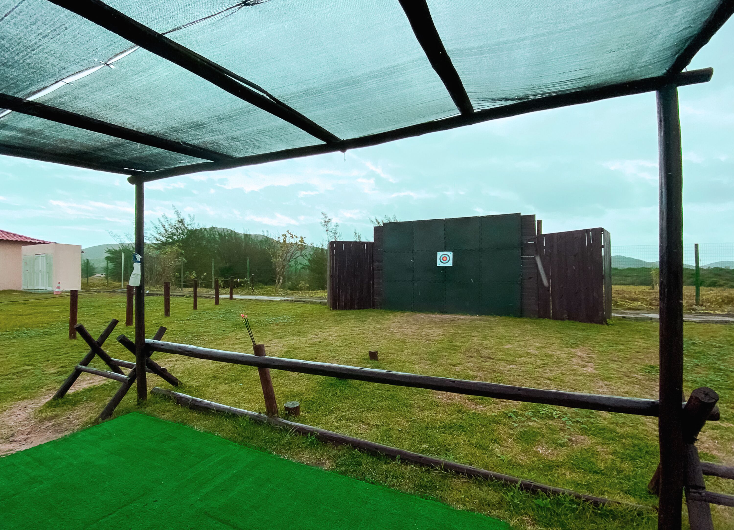 Un área exterior cercada con un objetivo para practicar tiro con arco, césped artificial verde y una estructura de madera al fondo.
