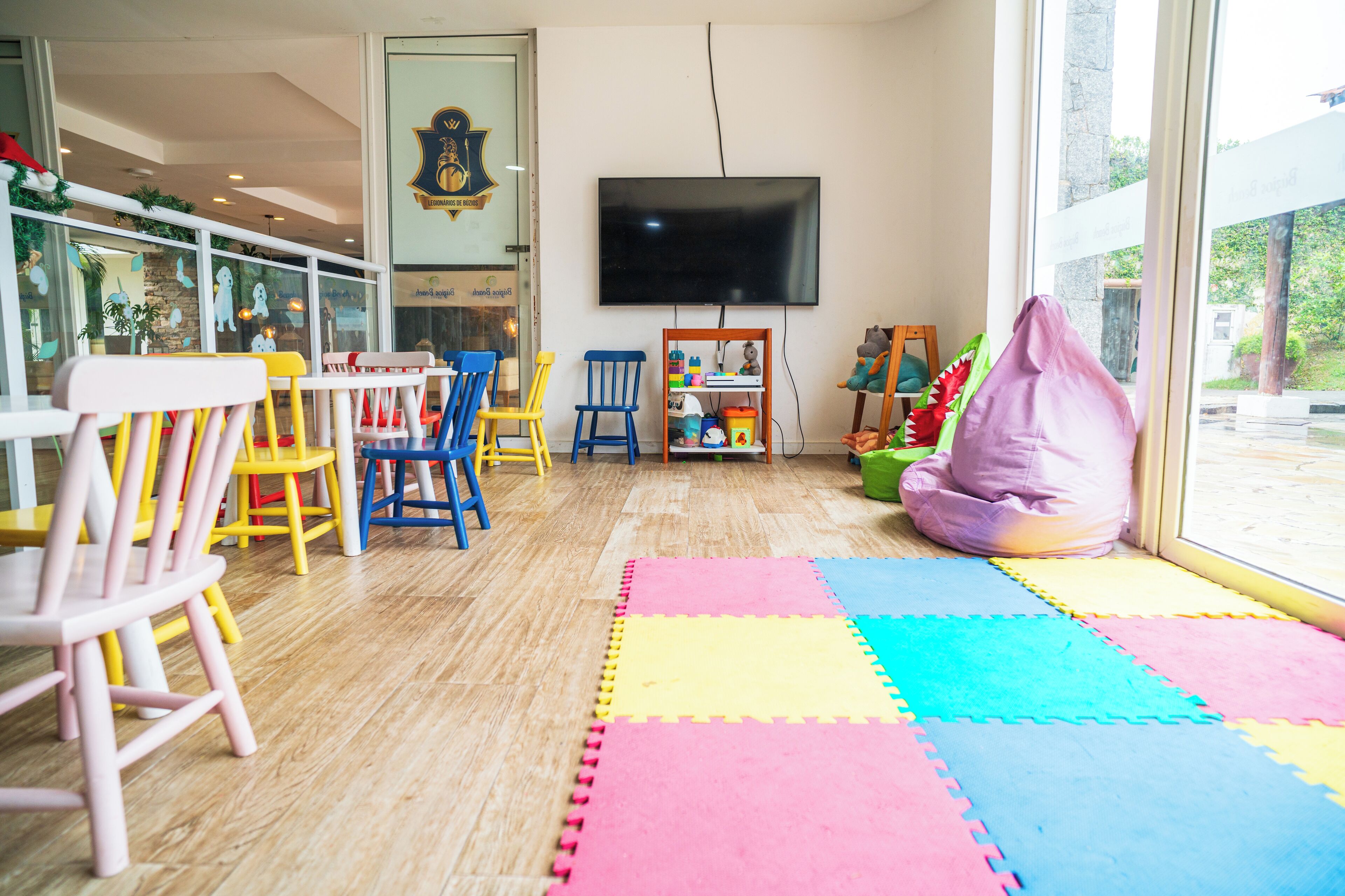 Un espace de jeux pour enfants avec des chaises colorées, un grand téléviseur et une fenêtre donnant sur un paysage verdoyant.