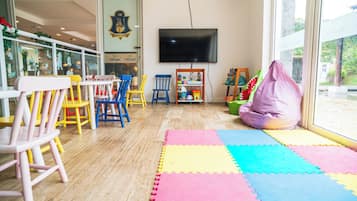 Un área de juegos para niños con sillas coloridas, un televisor grande y una ventana con vistas a follaje.