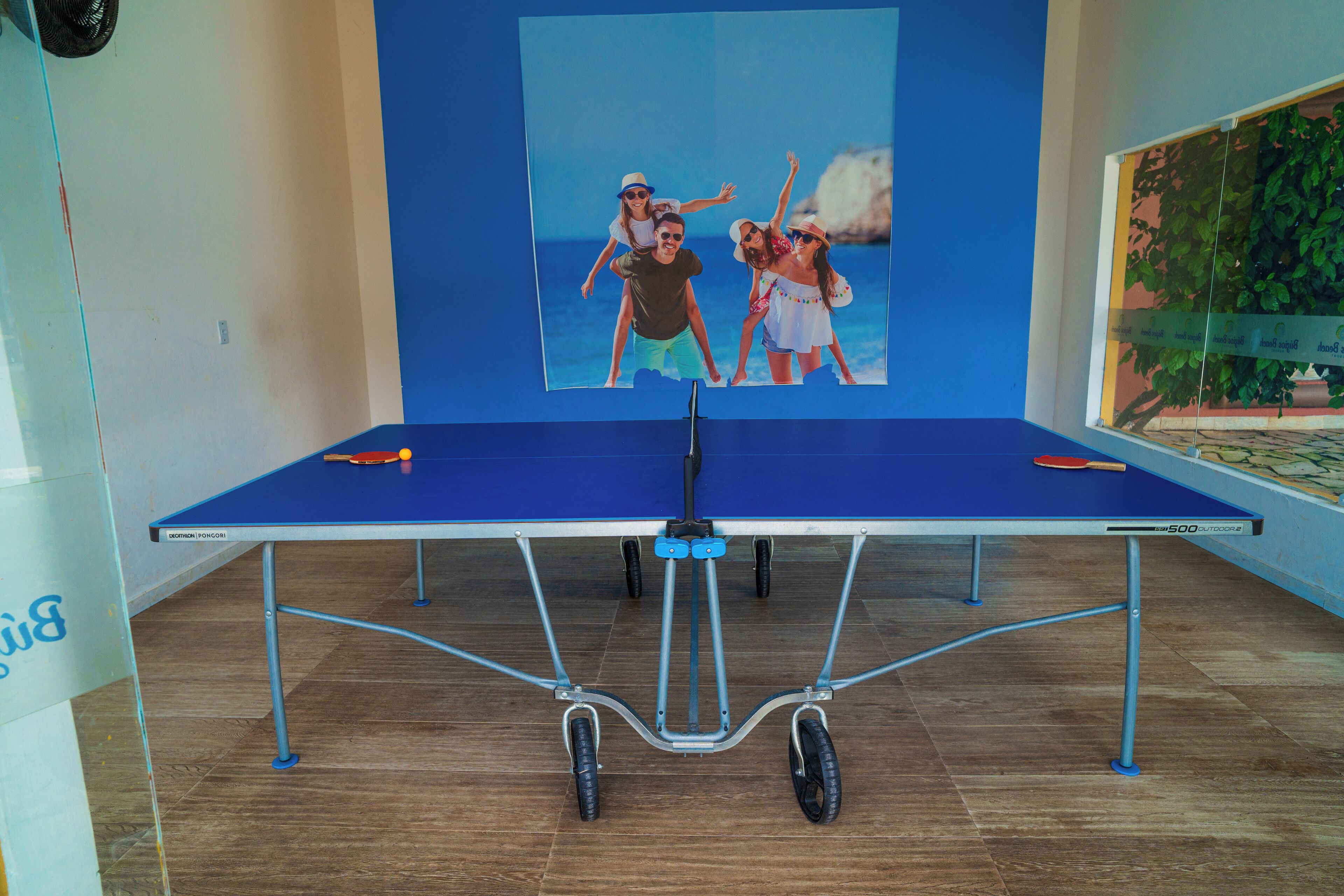 Una mesa de ping pong con mantel azul, un cartel con una familia en la pared y una planta en maceta a la derecha.