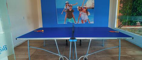 Una mesa de ping pong con mantel azul, un cartel con una familia en la pared y una planta en maceta a la derecha.