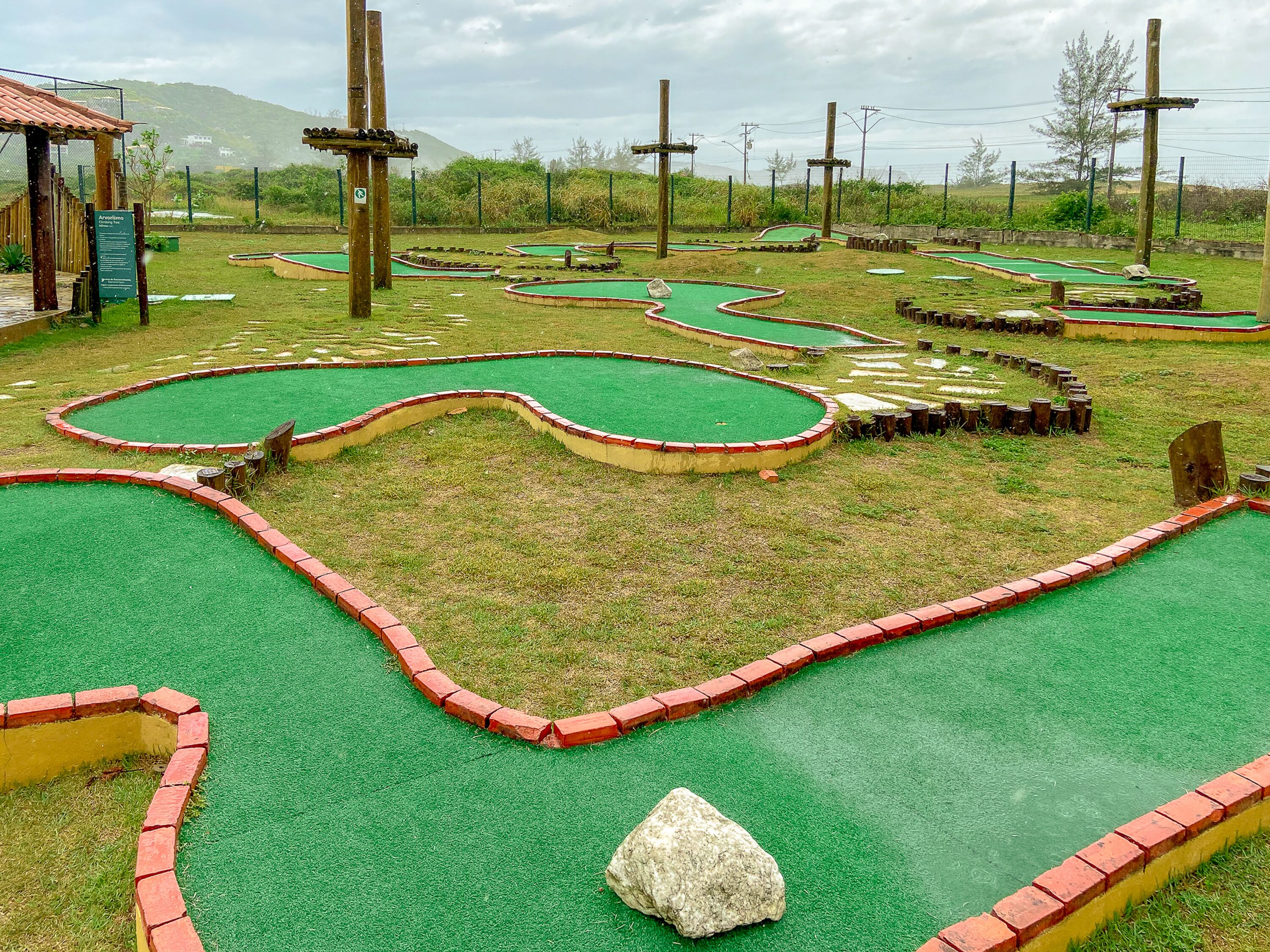 A miniature golf course with multiple green putting greens, red brick borders, and wooden structures.