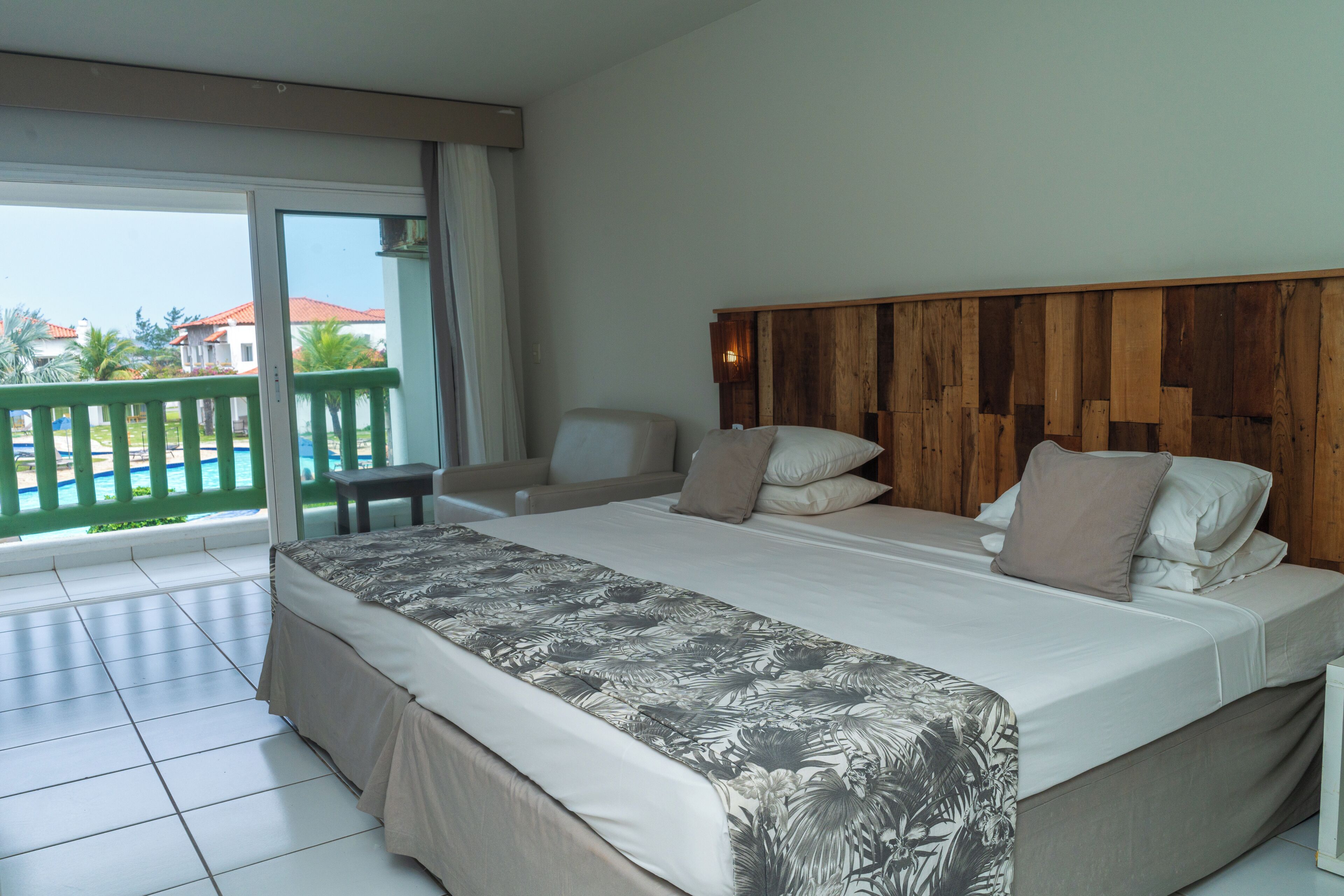 A hotel room with a large bed, a wooden headboard, a balcony with a view of palm trees and houses, and a chair near the window.
