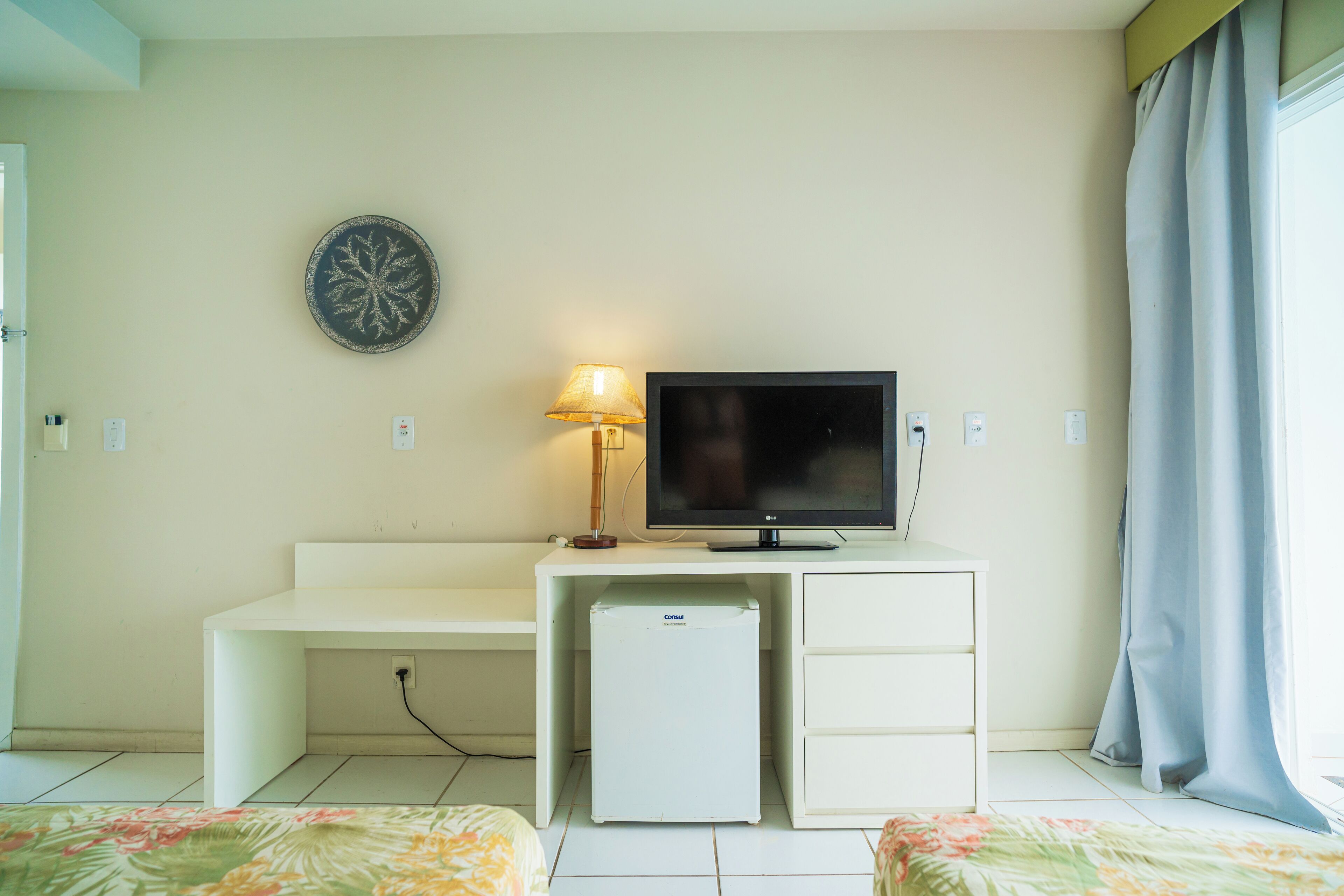 A hotel room with a bed, a desk with a television, a lamp, and a small refrigerator.