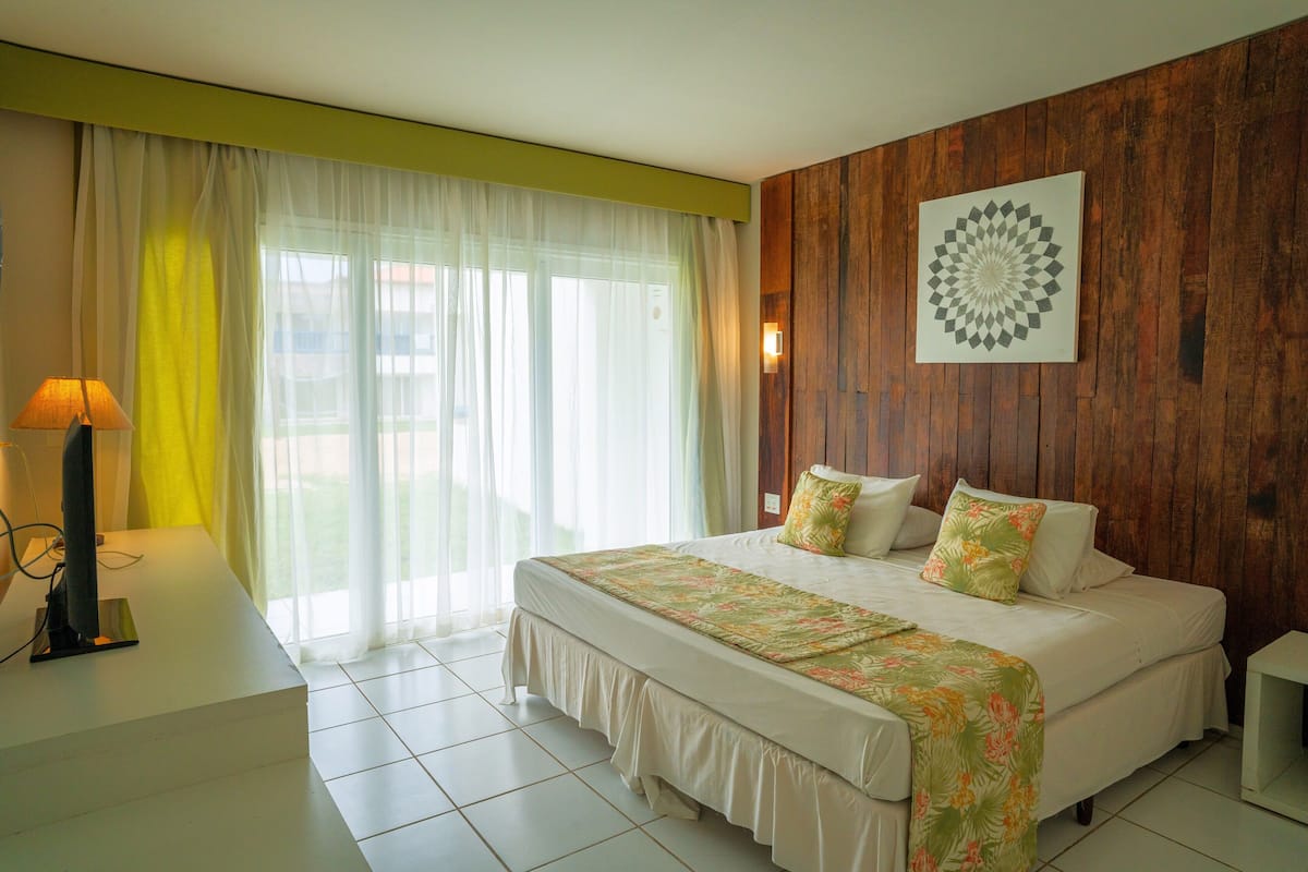 A bedroom with a large bed, a desk, and a window with sheer curtains.