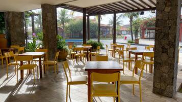 Un café al aire libre con sillas y mesas amarillas, pilares de piedra y vistas a una piscina y palmeras.